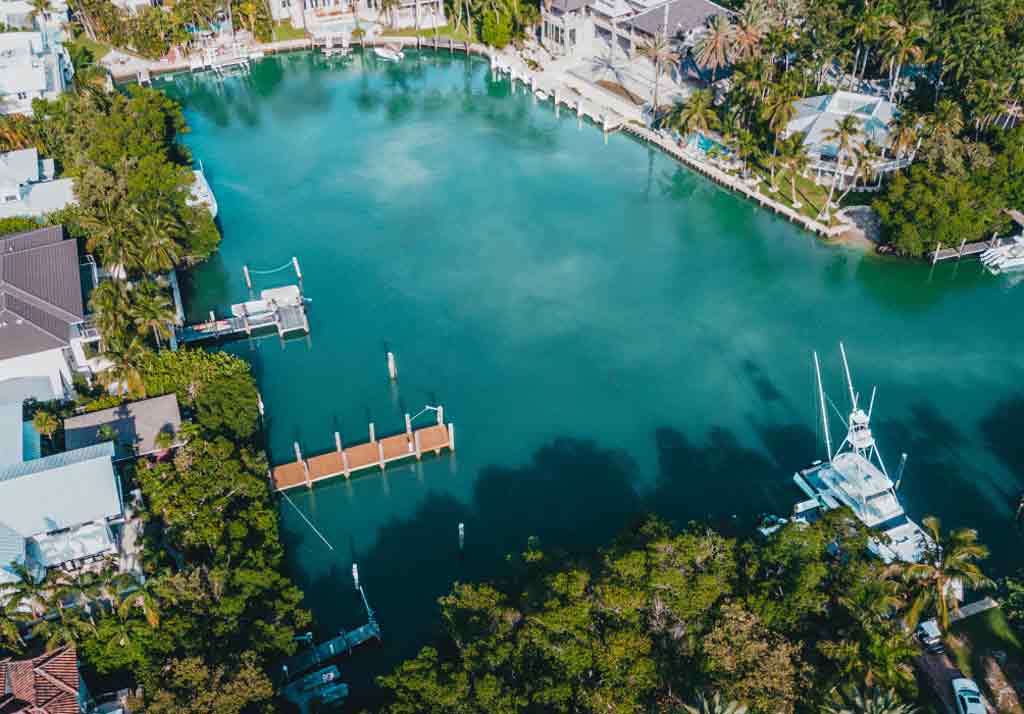 Aerial view of a serene residential waterfront neighborhood featuring a network of canals bordered by houses with private docks. Several boats are moored along the docks, and the homes are surrounded by well-maintained greenery, palm trees, and manicured lawns. The water reflects a calm blue-green hue, enhancing the tropical and tranquil atmosphere of the area. The scene suggests an affluent coastal community designed for boating and leisure.