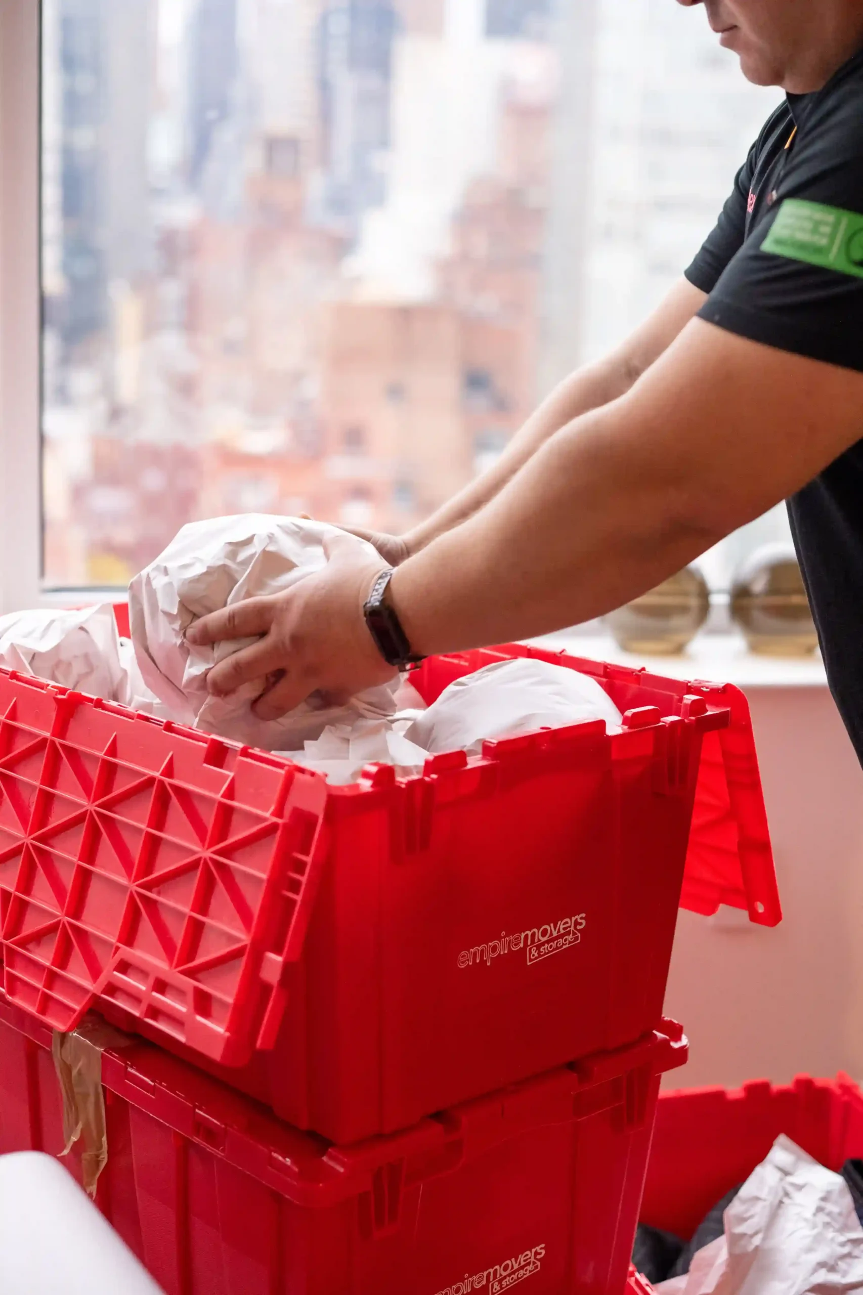 A person in dark clothing packs items into red plastic moving crates labeled with the “empiremovers & storage” branding. They are situated near a large window with natural light streaming in. Outside the window, a city skyline with high-rise buildings is visible. The atmosphere suggests an urban residential or office move.