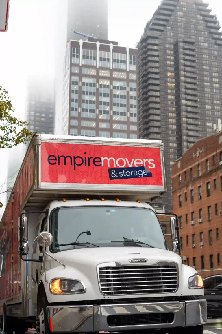 A moving truck branded “Empire Movers & Storage” is parked on a city street surrounded by tall, modern buildings. The truck is white with red and blue graphics, including the company name, phone number, and website. Fog partially obscures the upper sections of the skyscrapers, adding a moody urban atmosphere to the scene.