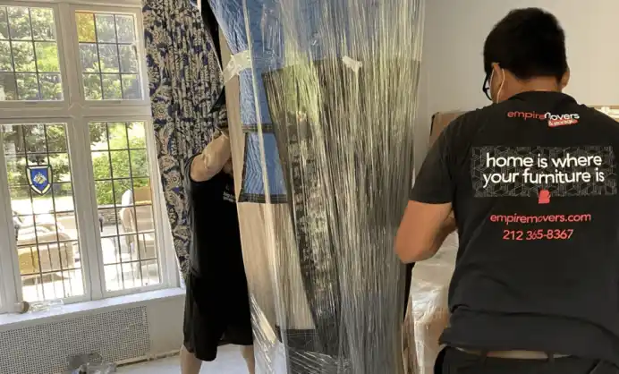 Two movers carry a large piece of furniture wrapped in clear protective plastic through a room with patterned curtains and a garden view visible through expansive windows. One mover wears a black t-shirt with the “Empire Movers” logo and slogan: “home is where your furniture is,” along with the company’s website and phone number. The other person, wearing a light-colored shirt, is partially obscured. Packing materials are visible in the room, suggesting an active moving process.