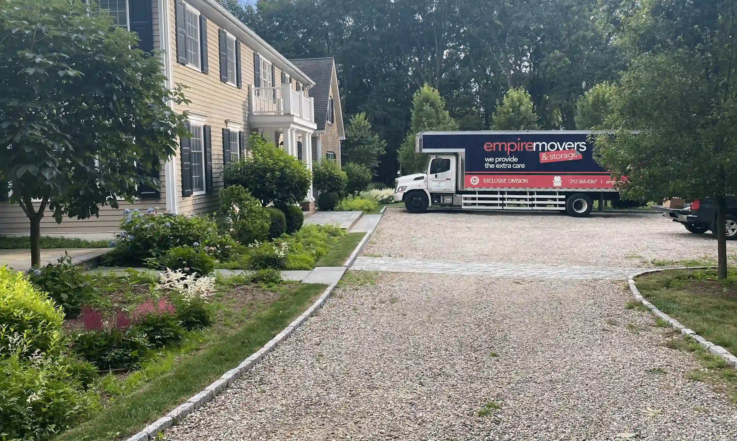 Movers NYC to DC. A moving truck from “Empire Movers & Storage” is parked on a gravel driveway in front of a large beige suburban home. The truck is white with blue and red graphics, displaying the company name, slogan “we provide the extra care,” phone number “212-365-8367,” and website “goempiremovers.com.” The house features black shutters, white trim, and a small balcony, surrounded by lush greenery and a manicured garden.