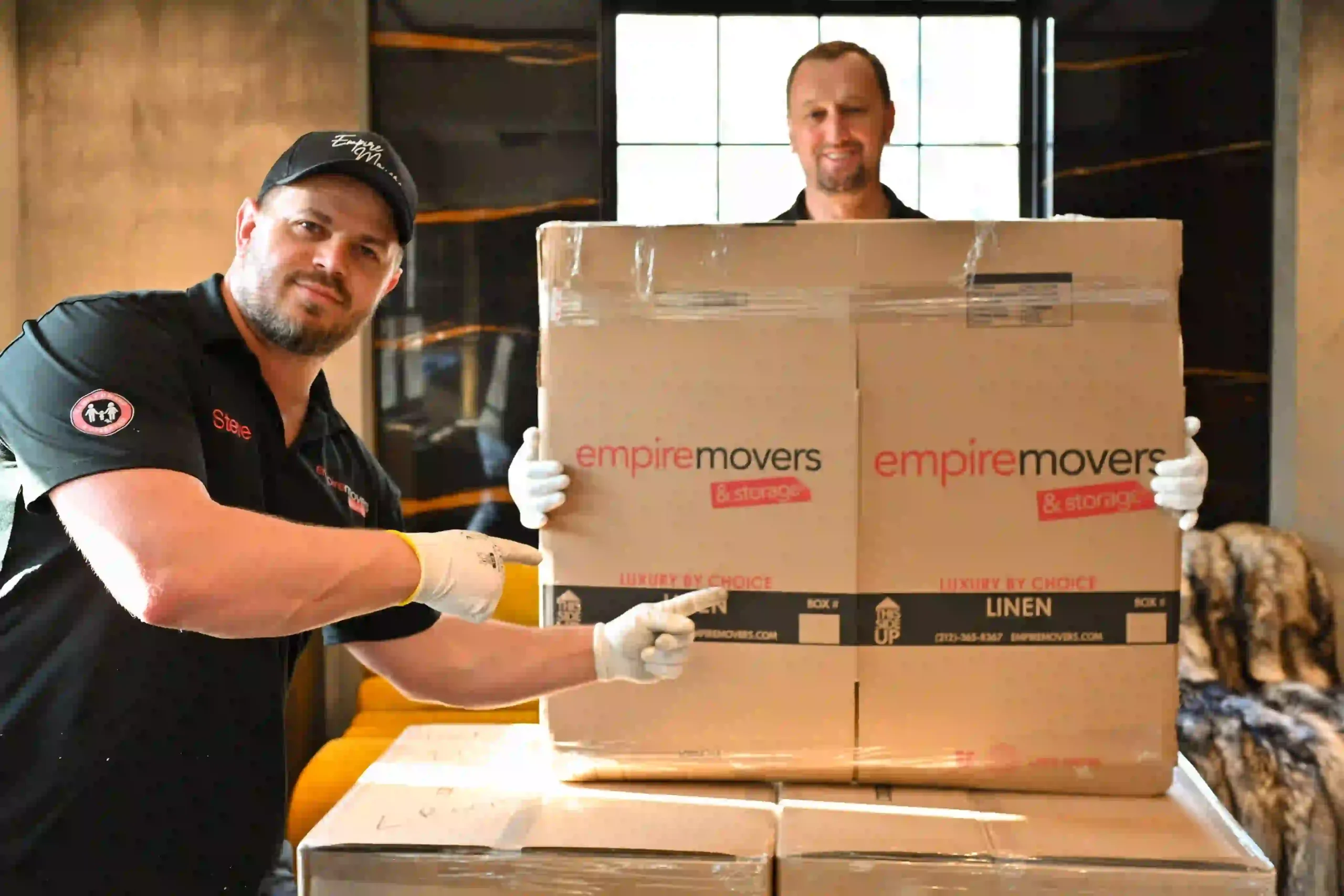 Two professional Empire Movers and Storage NYC crew members wearing uniforms and white gloves holding a large branded moving box marked 'Linen' in a luxury apartment setting.